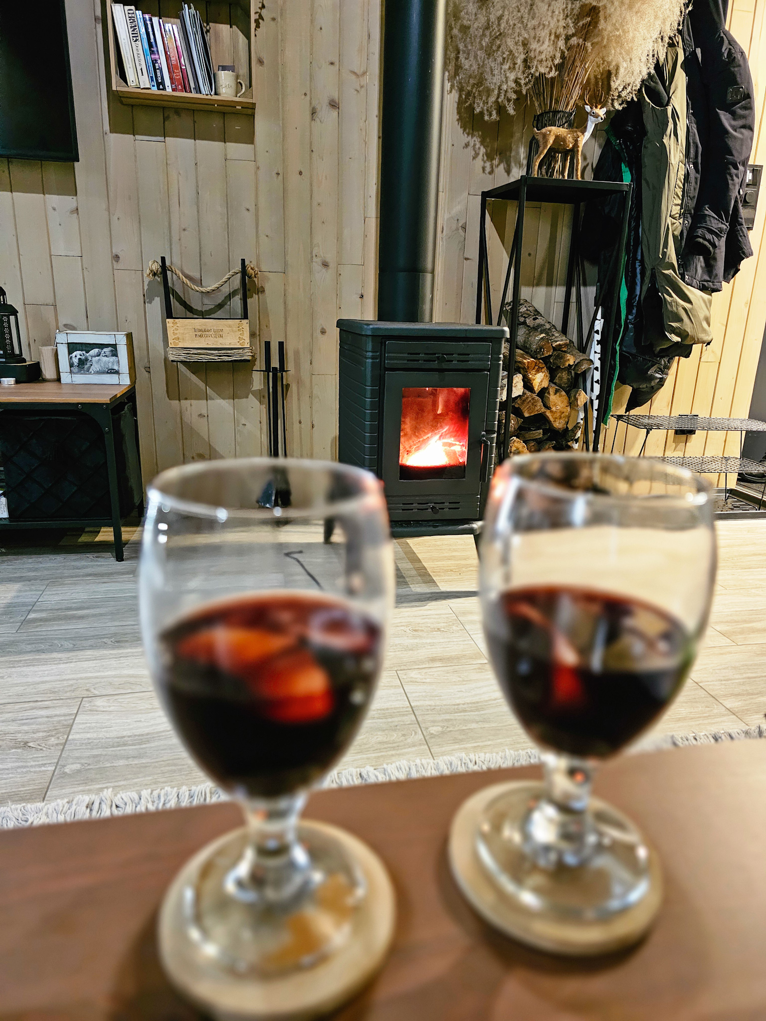 Dwa kieliszki z grzanym winem stoją na drewnianym stoliku. W tle widać przytulne wnętrze z kominkiem, w którym pali się ogień, drewnianymi ścianami oraz półką z książkami. Obok kominka ułożone są szczapy drewna, a na metalowym stojaku wiszą kurtki.