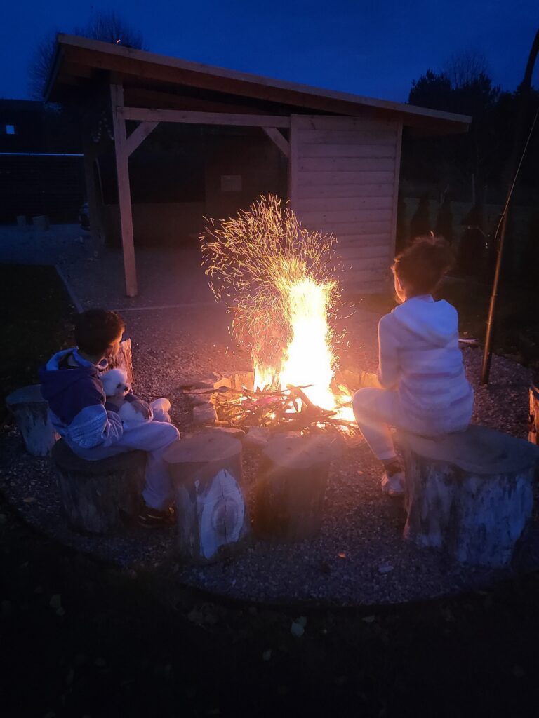 Dwójka dzieci siedzi przy ognisku za domkiem Hytte na Wyspie, ogrzewając się w ciepłym blasku płomieni. Wieczorny relaks na łonie natury to doskonały sposób na rodzinny wypoczynek nad Zalewem Nielisz. Cisza, spokój i magia ogniska tworzą niezapomnianą atmosferę.